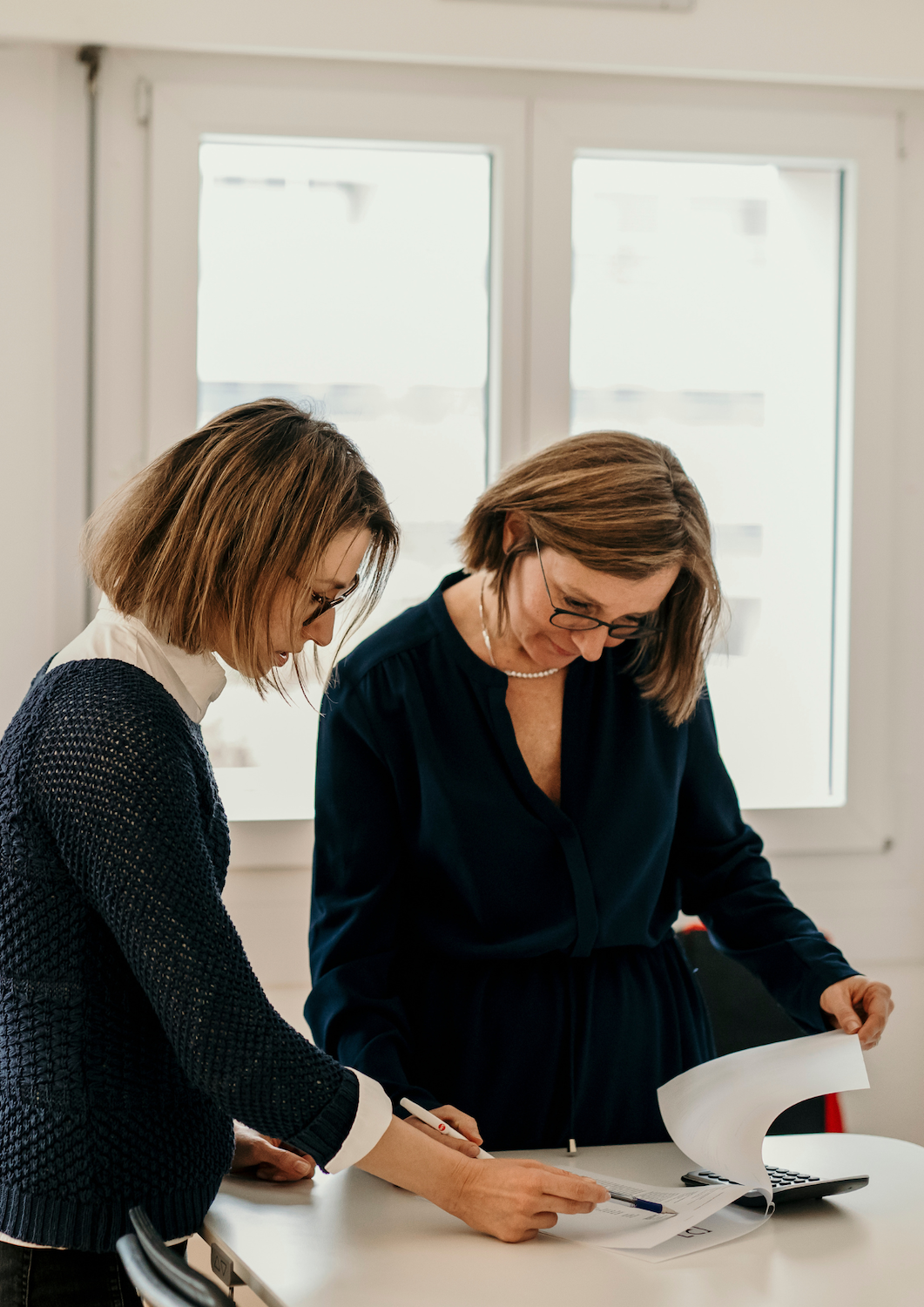 Deux femmes habillées de manière professionnelle, dont Kathleen Druart (fondatrice d’Enfin!Fidu), discutent autour de documents stratégiques sur un bureau, illustrant le partenariat humain et l’analyse sur-mesure au service de la croissance d’une PME.