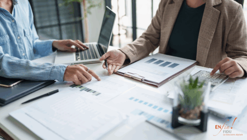 Deux professionnels concentrés autour d’un bureau avec ordinateur, calculatrice, graphiques imprimés et classeurs, illustrant une session d’analyse financière dans une PME.