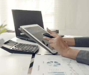 Gestion financière moderne pour PME - EnfinFidu Femme assise à un bureau utilisant un iPad avec calculatrice, graphiques et ordinateur pour analyser la situation financière de son entreprise