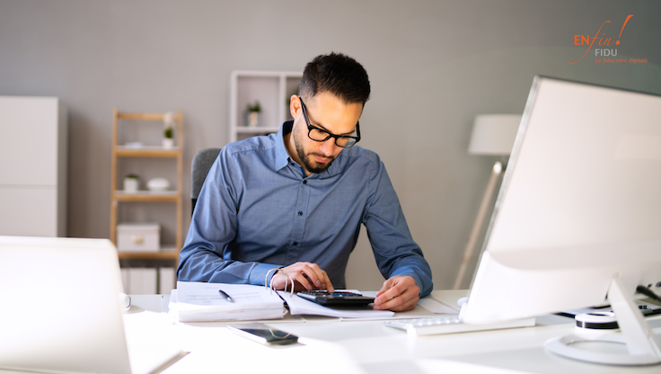 Homme d’affaires assis à son bureau, calculette à la main, analysant des documents financiers sur ordinateur pour automatiser la comptabilité de sa PME - EnfinFidu